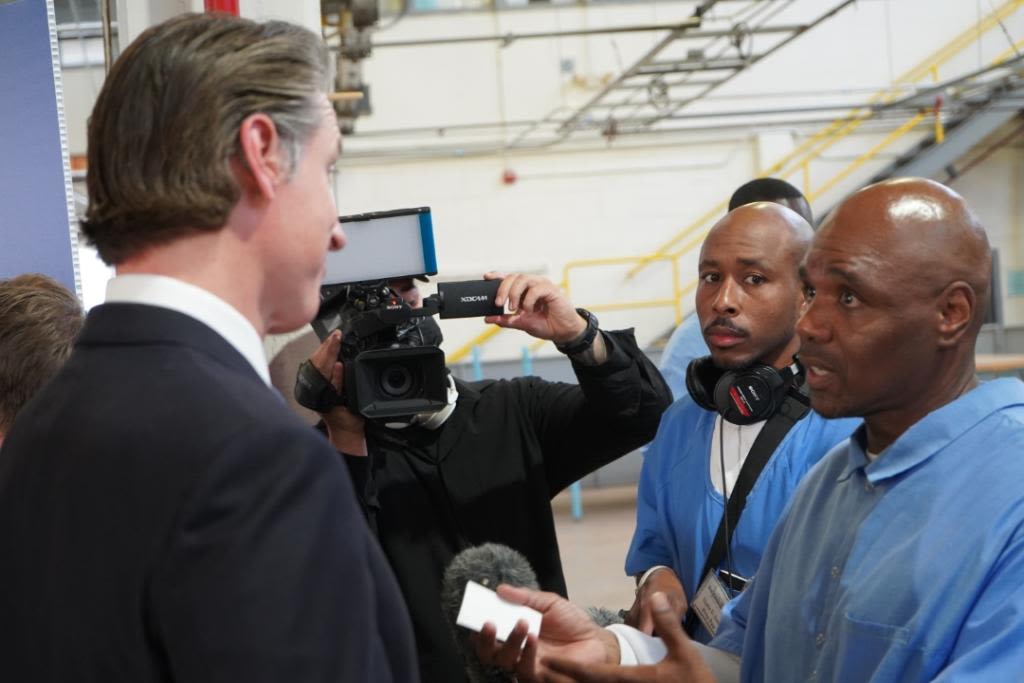 Governor Gavin Newsome talks with two men in blue prison jumpsuits, one of whom is Steve Brooks. Another person films. 