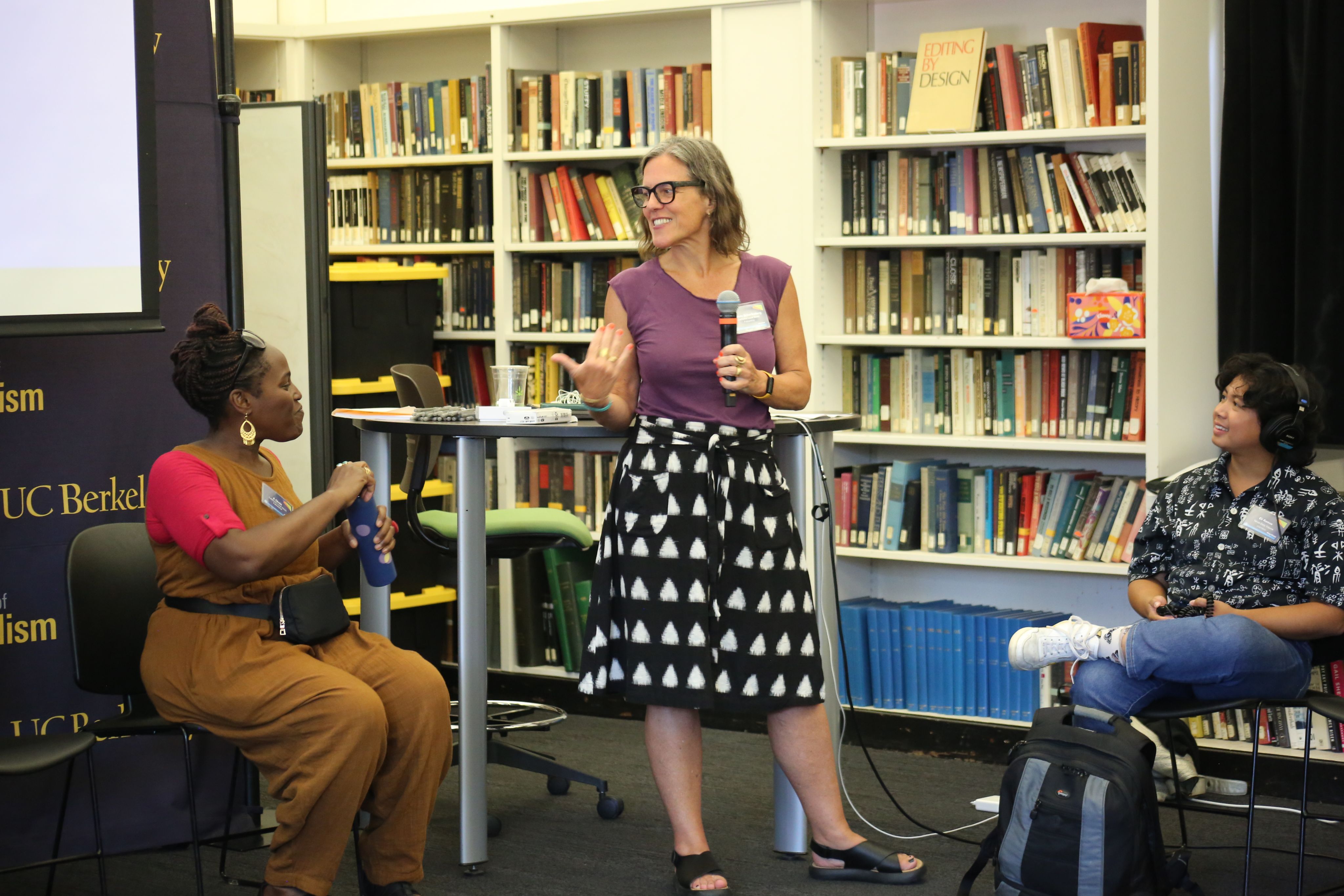 Christa Scharfenberg in a polka dot skirt holds a mic and gestures while talking to a colleague who is seated. A fellow in the program looks on.