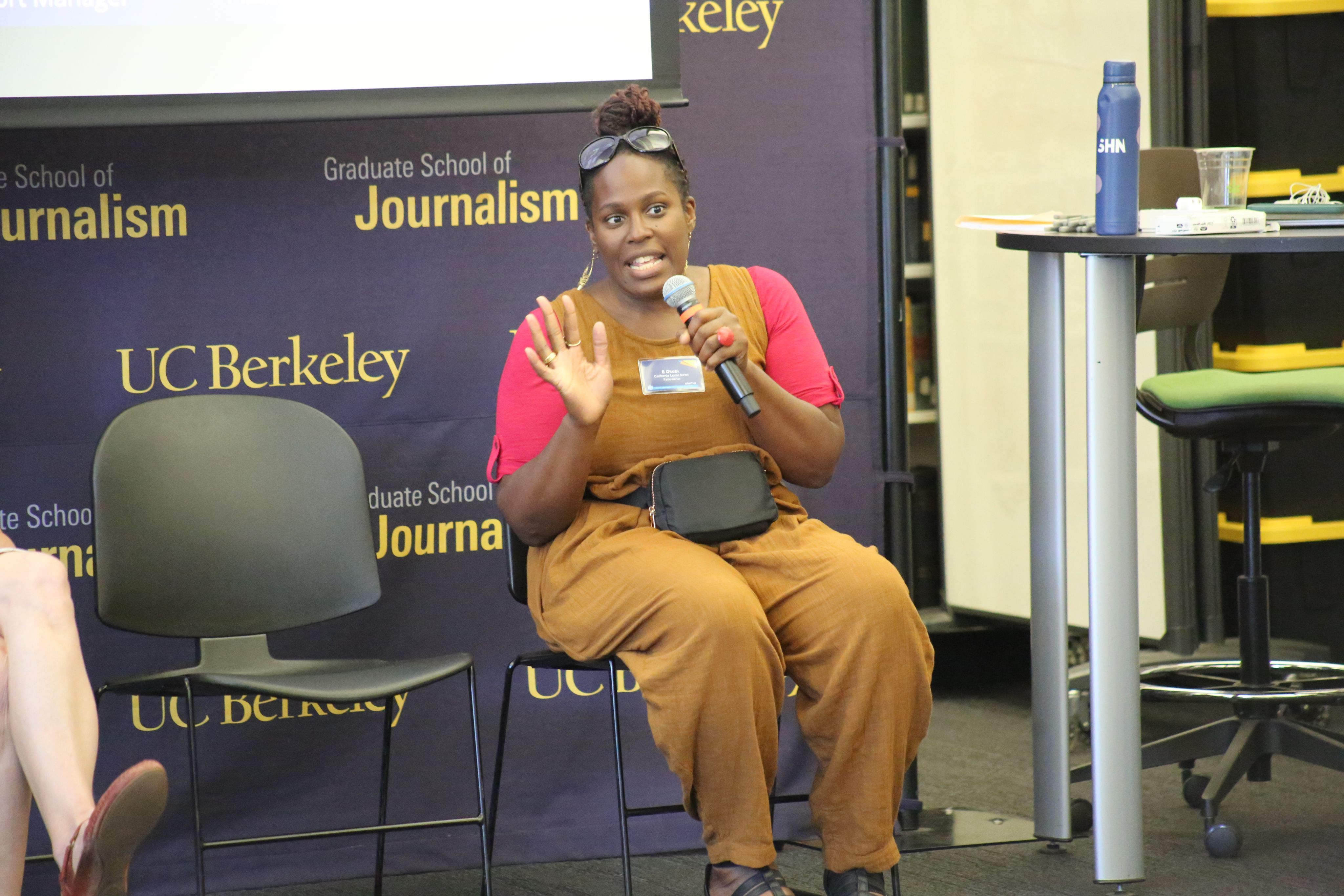 A woman in  brown coveralls and a pink shirt speaks into the camera with one hand raised. 