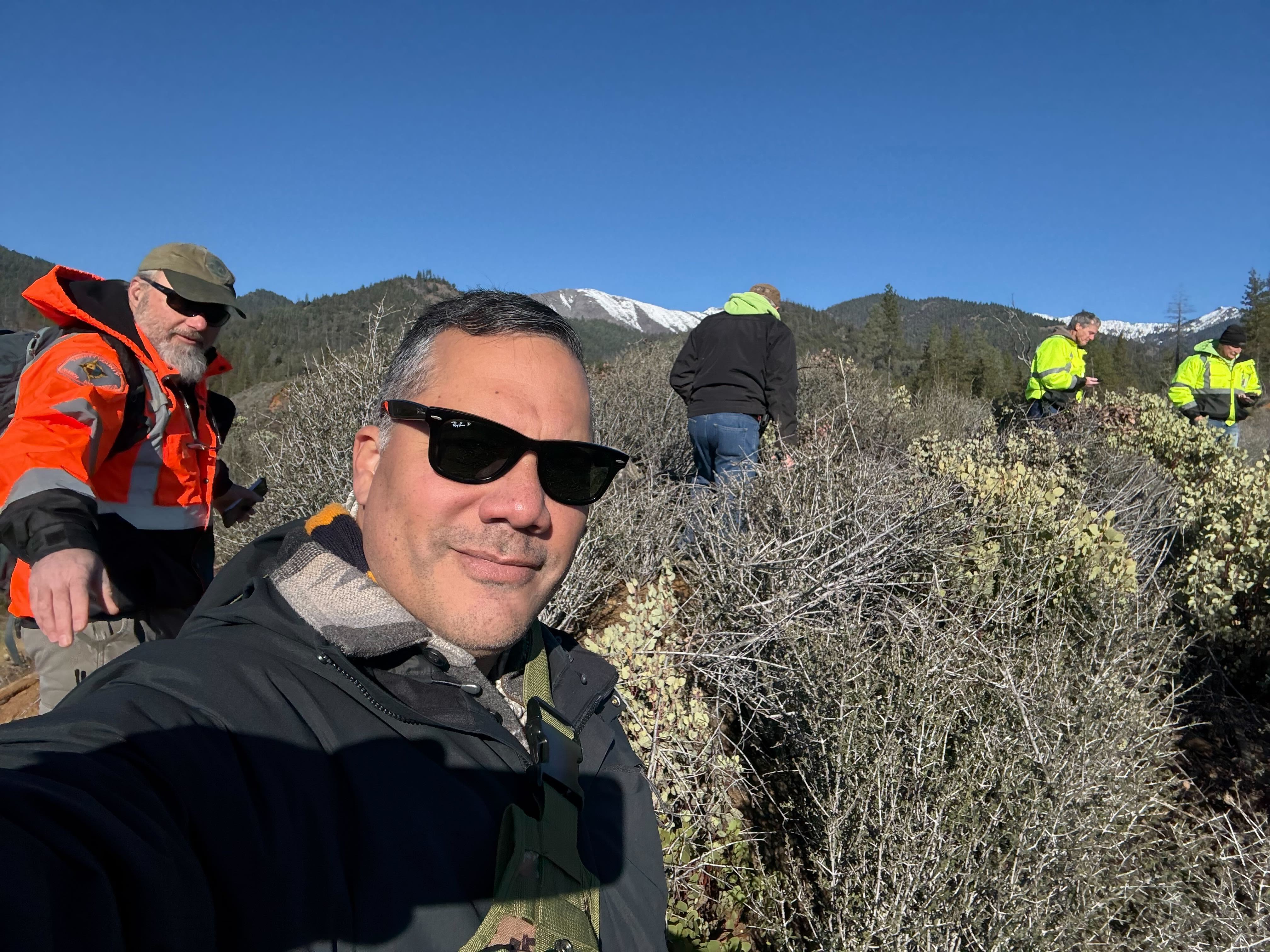 close up of a man taking a selfie with people in orange and yellow jackets in the brush outside of a mountain range.