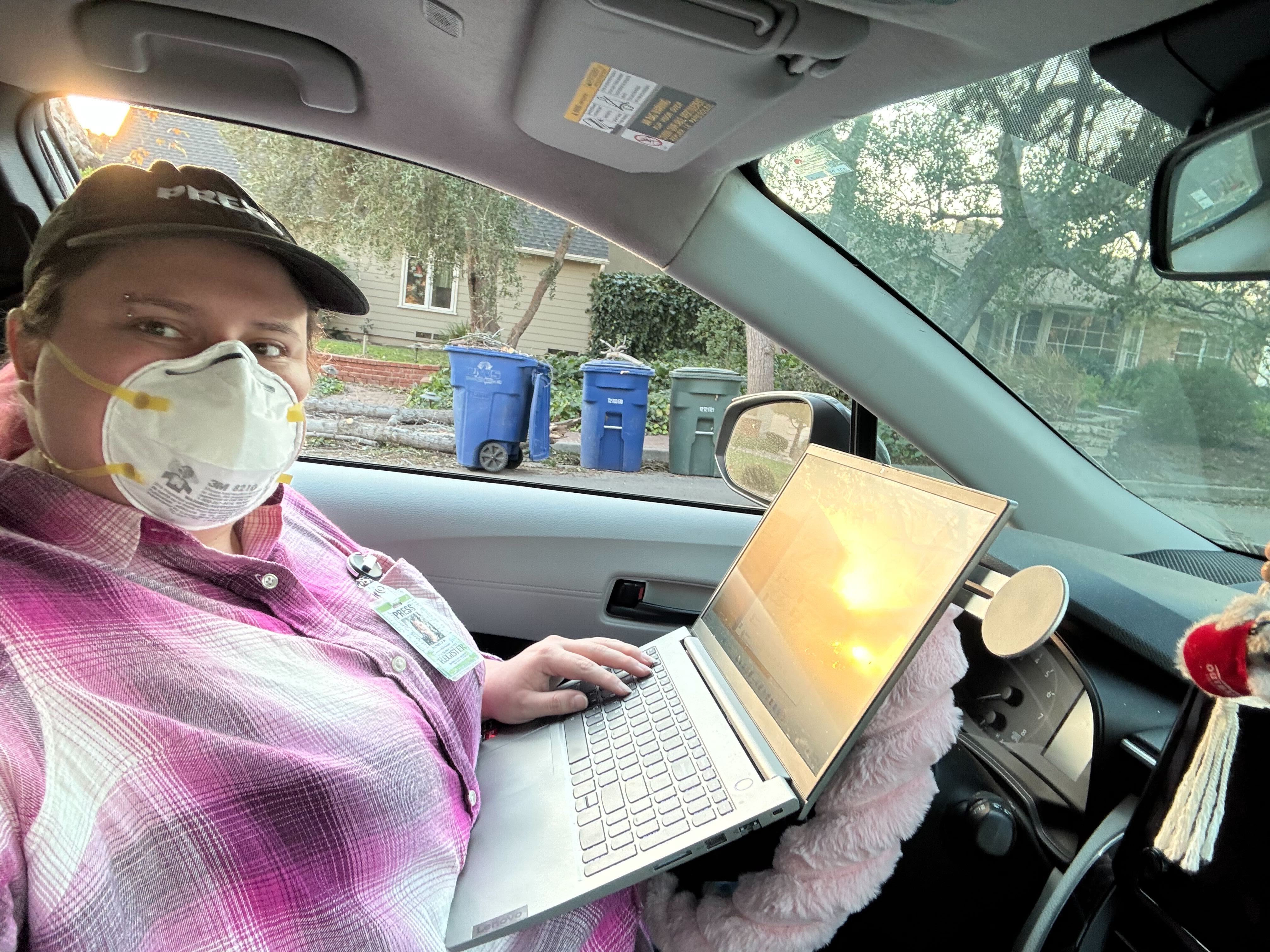 a person wearing a baseball cap and a mask in their car with a computer on their lap. An image of fires is on the computer screen.