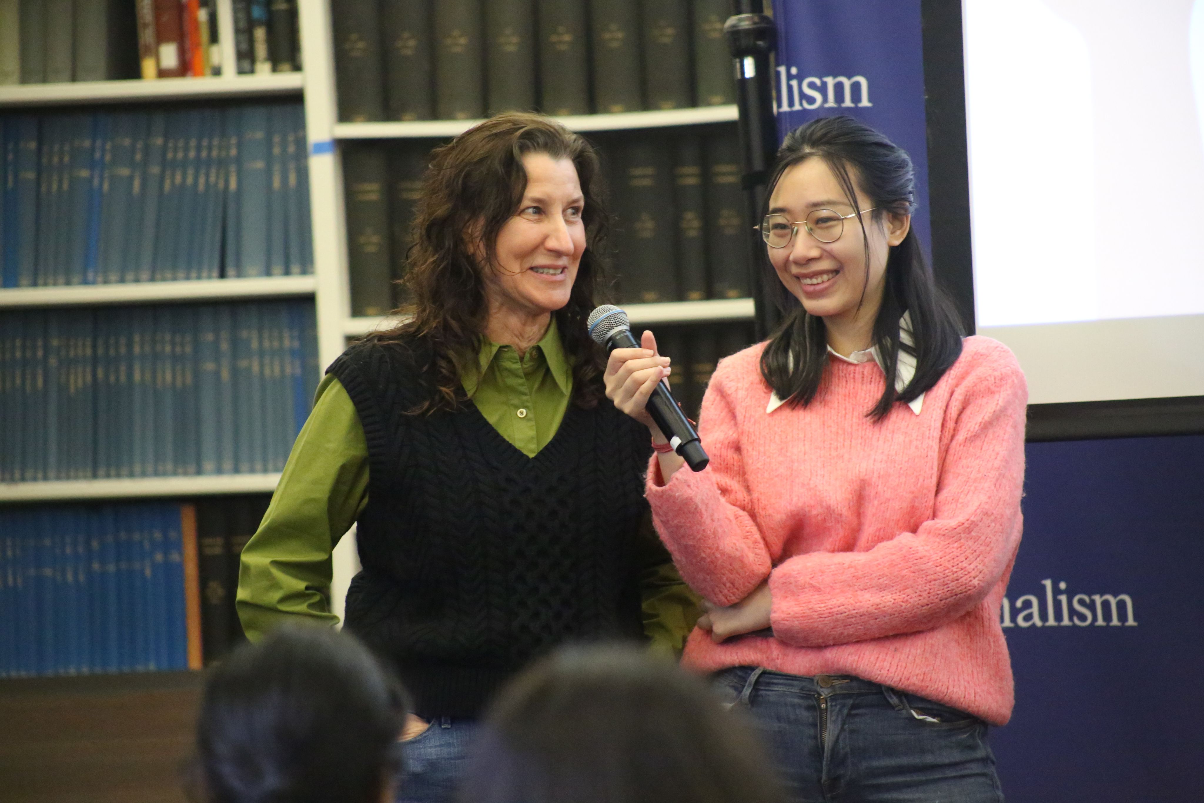 Two women speaking into one mic, one in a tree green shirt and black vest and another in a pink sweater. They are smiling.