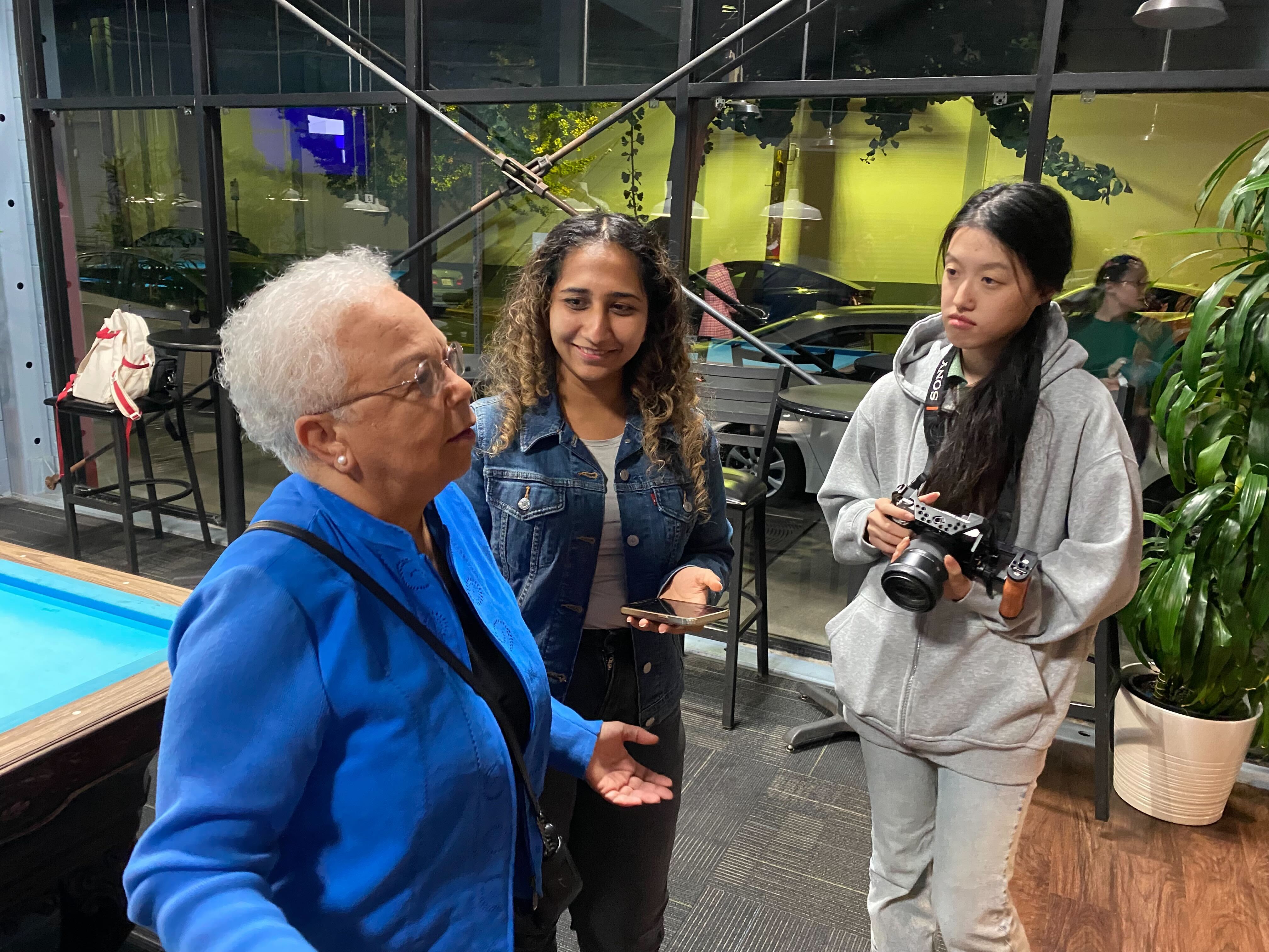 Two students, one with a cell phone and another with a camera, talk with an older woman in a blue jacket. 