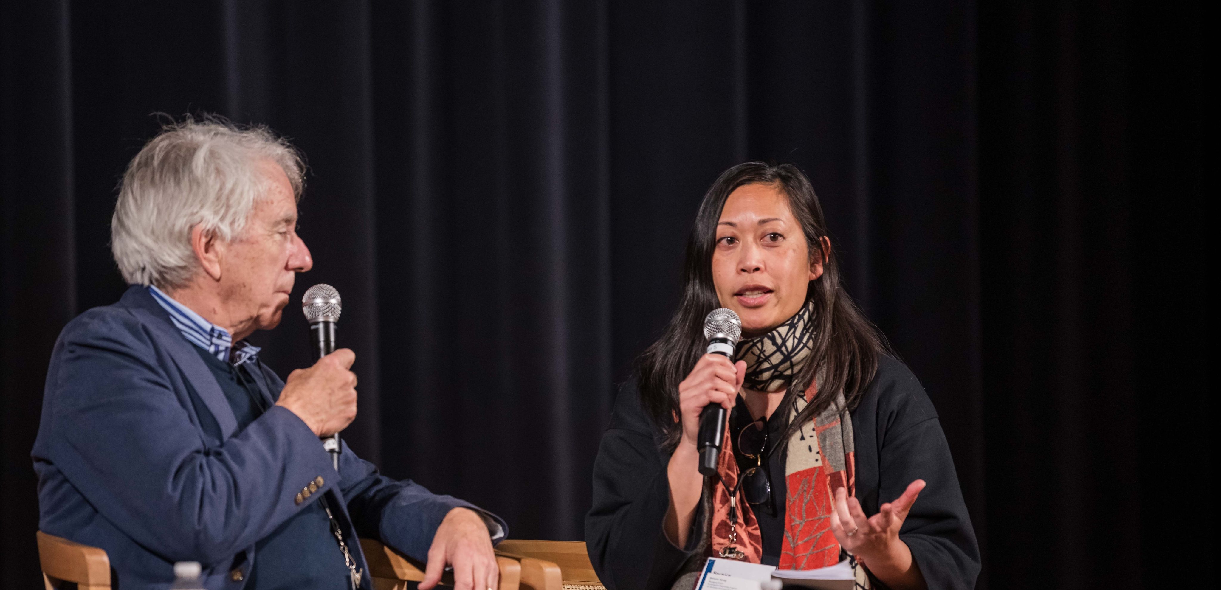 A woman with a microphone and colorful scarf talks with a man in a blue suit holding a mic.