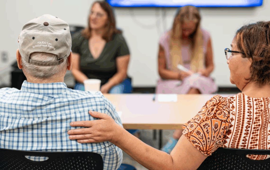 A woman with her hand on the back of an older man sitting around a table of people talking. 