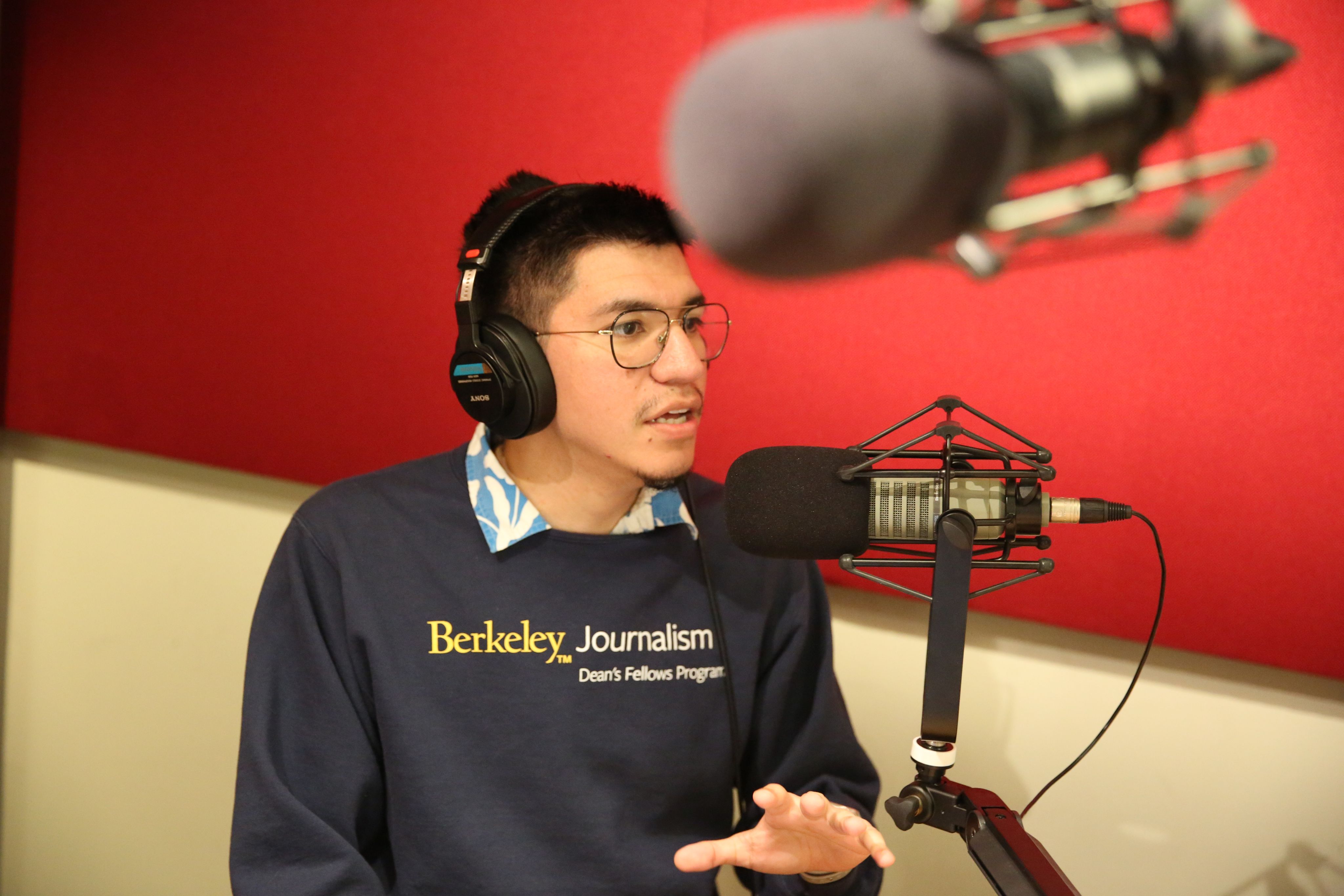 Andres Larios wears a Berkeley Journalism sweatshirt in the school's radio lab. You can see the microphone in front o f him and a red wall behind. 