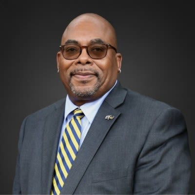 Headshot of Dean Michael Bolden wearing a yellow and blue striped tie and suit jacket. He is wearing glasses. 