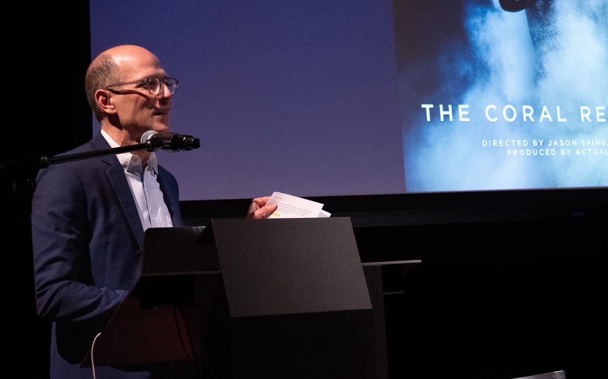 Jason Spingard Koff speaks at the Ocean's Stories symposium. He is wearing a blue sports coat and a microphone is in front of him.