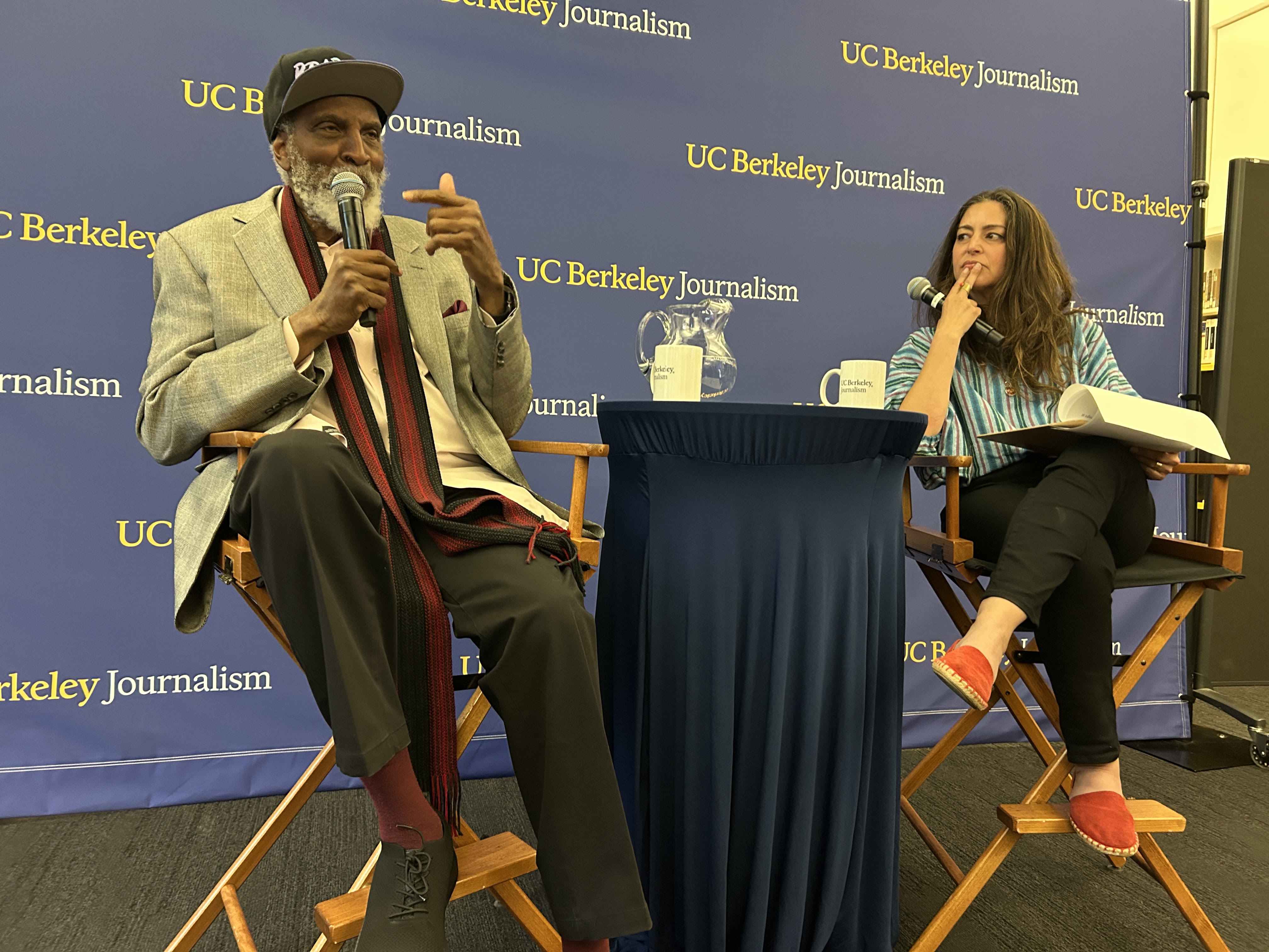 Professor john powell is on stage in front of the UC Berkeley Journalism banner with Shereen Madison Meraji. 