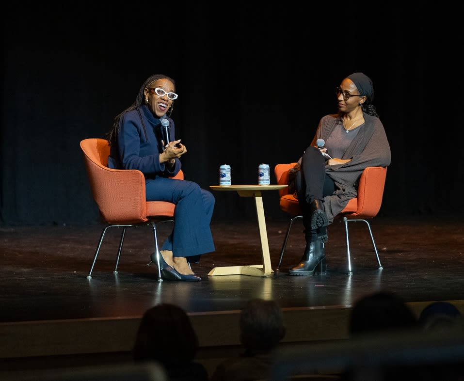 Two women with microphones with a black background sit on stage with orange chairs. 