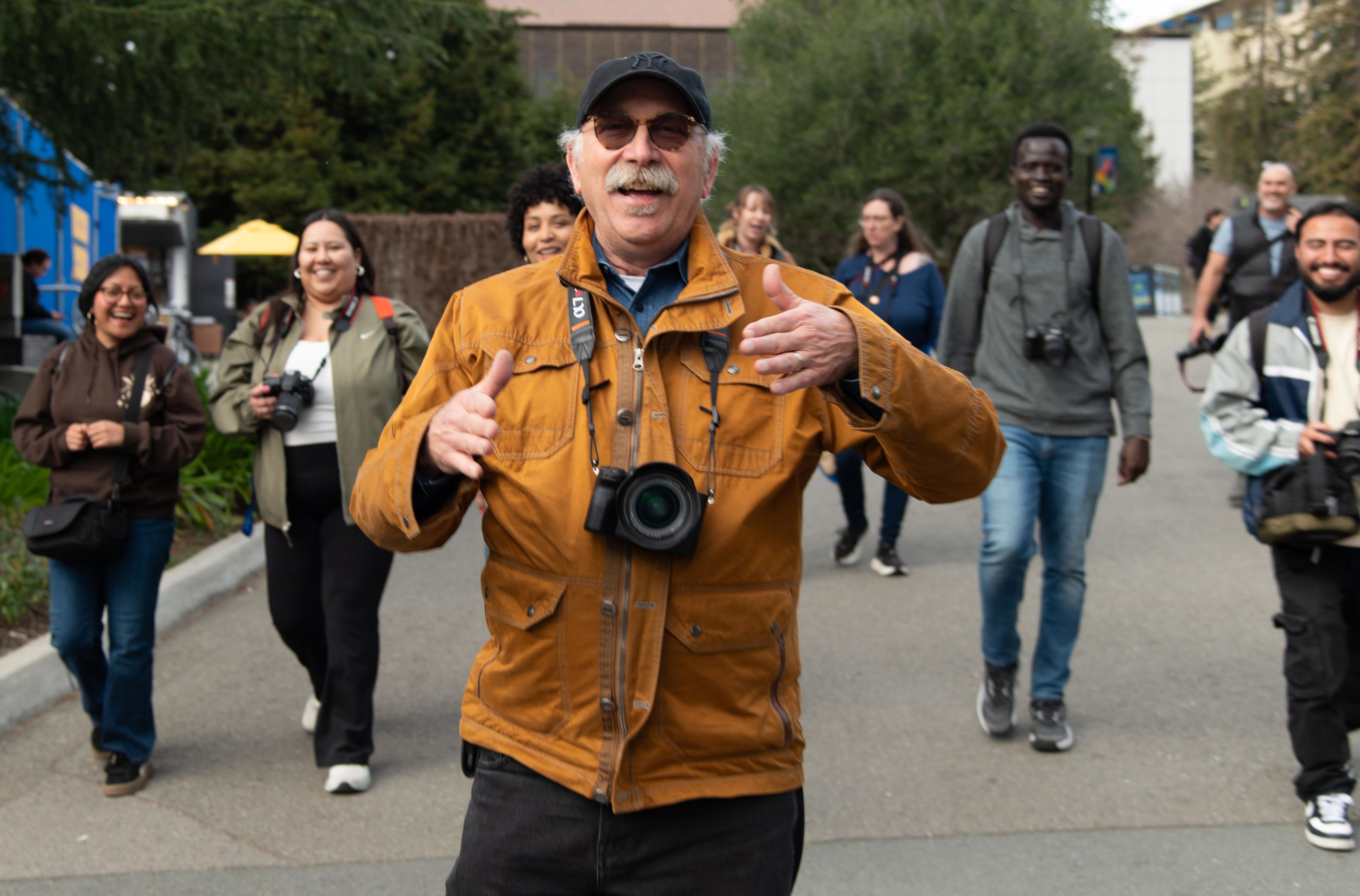 Ken Light is wearing a yellow/orange jacket and walking in front of his students, who are smiling. 