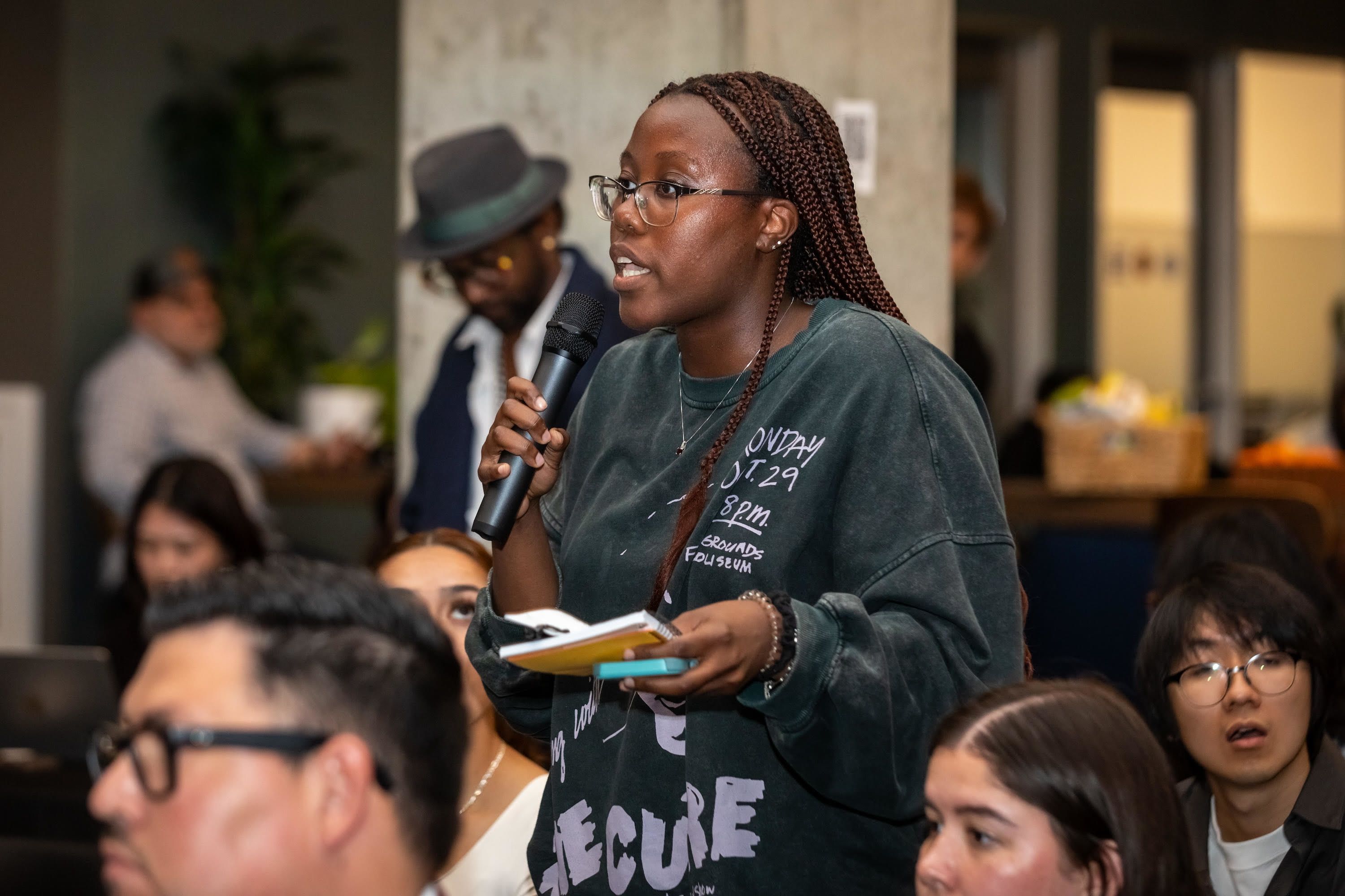 Skylla Mumana speaks with a microphone and holding a notepad during a debate in Richmond. She is asking questions from someone on the stage.